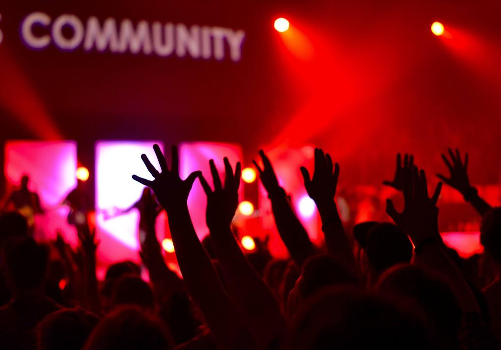 Man sieht die Hände einer feiernden Menschenmenge im rotem Licht. Das Wort 'Community' wird verschwommen an die Wand darüber projiziert.